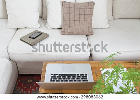 Still life detail of luxury living room with white sofa and coffee table with open laptop computer and smartphone with paperwork and folders, home interior. Aspirational technology indoors lifestyle.