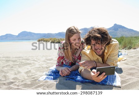 Portrait of a smiling young tourist couple using technology to take selfies photos, enjoying a summer holiday together, laying on a sandy beach on vacation, outdoors. Travel and technology lifestyle.