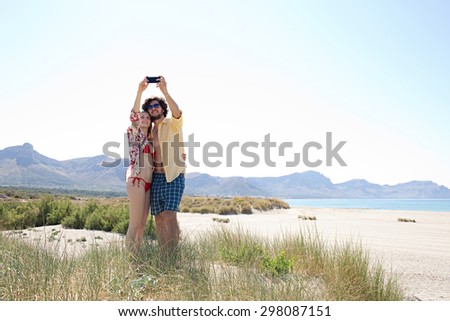 Young tourist couple on beach with dunes and mountains together, enjoying a sunny summer holiday, using a smartphone to take selfies, networking outdoors. Recreational travel lifestyle.