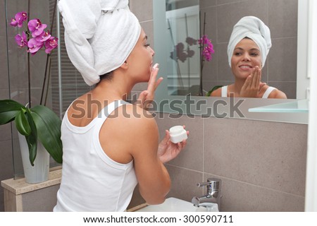 Side portrait and face reflection of a joyful young woman applying nourishing cream on her face, looking at herself in a bathroom mirror, with towel wrapping her hair, home interior. Skin care.