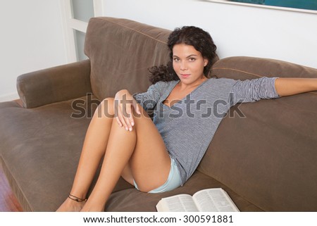 Portrait of beautiful young woman lounging on sofa at home reading a book, sitting relaxing in living room interior. Relaxing home hobbies lifestyle, indoors. Girl reading a book at the weekend.