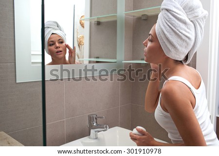 Side portrait and face reflection of a beautiful young woman applying nourishing cream on her face, looking at herself in a bathroom mirror, with towel wrapping her hair, home interior. Skin care.