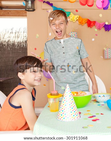 Two children brother and sister enjoying a birthday party at a celebratory fun table with food and sweets in a home garden, outdoors. Kids blowing party blowers, having fun together, home exterior.