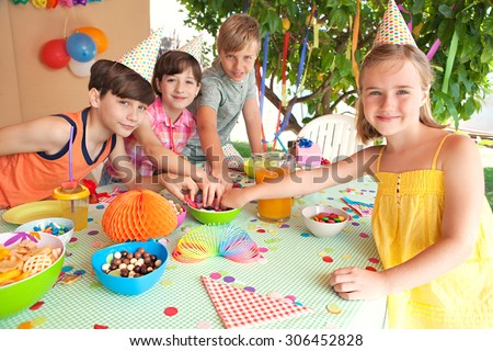 Group of children at a home garden birthday party having fun and eating sweets and party food, outdoors. Kids enjoying a party wearing paper hats and blowing party blowers on sunny day, lifestyle.