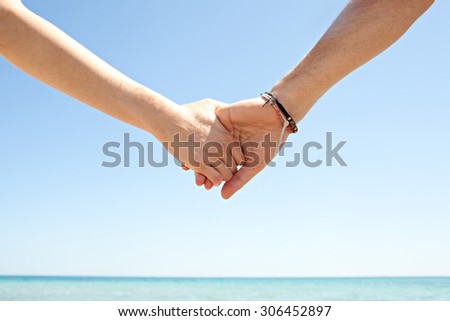 Detail view of a man and a woman hands being held together against a blue sky and sea, on a sunny summer holiday break, outdoors. Tourists holding hands, lifestyle. Honeymoon romance on beach.