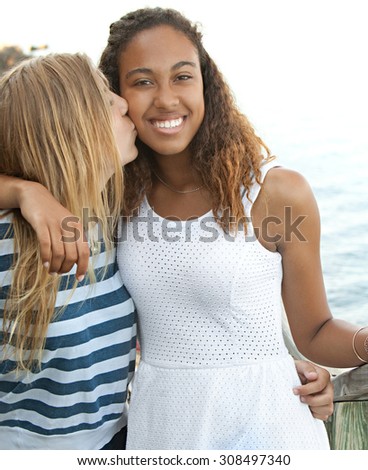 Beauty portrait of two teenager girls of different ethnic origins friends hugging and kissing, smiling at the camera by the sea on a summer holiday, outdoors. Travel lifestyle, nature beach exterior.