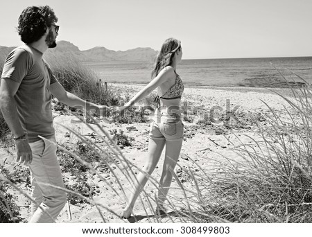 Black and white view of young tourist couple with woman taking man by the hand, walking towards the sea, enjoying a summer holiday together on a beach. Travel and lifestyle vacation, nature exterior.