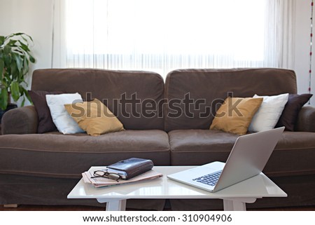 Still life of living room with open laptop computer, paperwork and spectacles in a family home with comfortable sofa by large bright window, house interior. Working from home with technology, indoors.