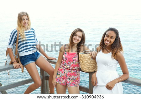 Diverse group of three teenager girls standing on a wooden deck together, smiling on a beach with a turquoise sea background. Adolescent girls spending holiday time together on a summer day, exterior.