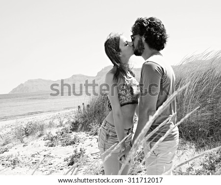 Black and white view of young tourist couple kissing on beach with clear waters and mountains, enjoying a summer holiday together on a nature exterior. Travel and lifestyle vacation, outdoors coast.