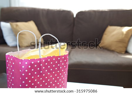 Close up detail still life of feminine paper shopping bag in a stylish bright living room, home interior. Pink carrier bag, indulgence and consumerism lifestyle in an elegant room with sofa, indoors.