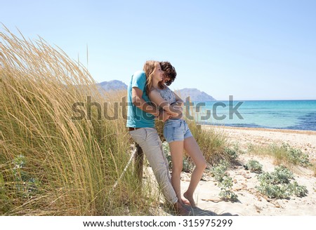 Attractive young tourist couple enjoying a summer holiday together on beach with blue waters and grass sand dunes, hugging and kissing in romantic summer honeymoon, outdoors nature. Travel lifestyle.