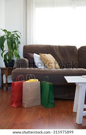 Still life view of paper shopping bags in a stylish bright living room, home interior. Carrier bags on a shiny wooden floor in an elegant room with sofa, indoors. Indulgence and consumerism lifestyle.