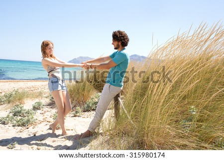 Joyful tourist couple enjoying a summer holiday together on beach with blue waters and grass sand dunes, holding hands being playful in a romantic summer honeymoon, outdoors nature. Travel lifestyle.