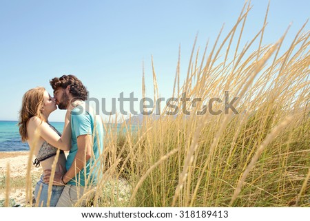 Attractive young tourist couple hugging and kissing on the lips standing on a beach with blue sky and long grass dunes on a summer holiday outdoors. Travel and honeymoon lifestyle, nature exterior.