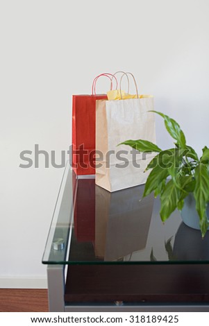 Two paper carrier shopping bags on a glass reflective table on plain white wall in home living room, interior. Consumerism and indulgence lifestyle indoors. Still life of shopping bags on office desk.
