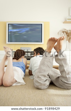Couple watching tv together while laying down on the floor in living room.