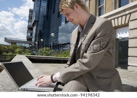 Young businessman typing on a laptop computer while sitting by modern office building, outdoors.