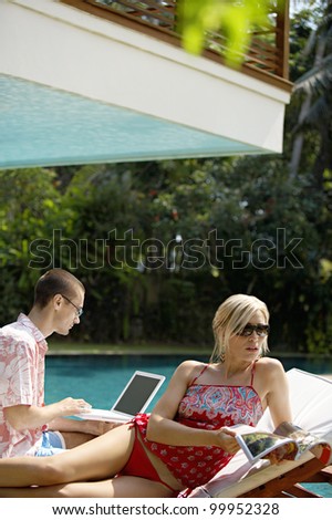 Young couple lounging by the pool in a garden, using technology and reading a magazine.