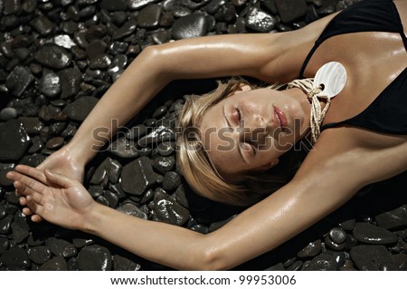 Over head view of a young woman wearing a black bikini and laying down on wet black stones with her eyes closed.