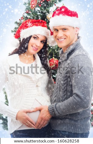 Young pregnant woman and happy father decorating Christmas tree over blue background with snow