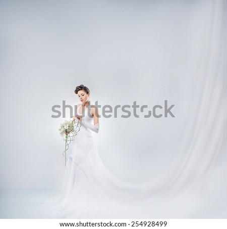 Young and beautiful bride in a long fantasy dress standing with a flower bouquet over the grey background.