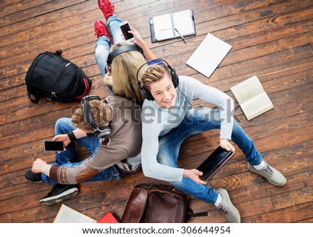 Group of students using smartphones and tablet in headphones listening to the music and leaning on each other on wooden floor having notebooks and bags around them.