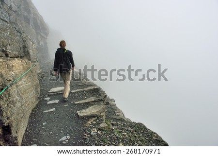 female hiker with a backpack on a steep highland alpine trail in heavy fog, view from the back