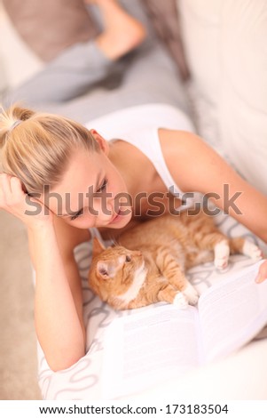 young woman lying on a sofa with her cat and reading a book