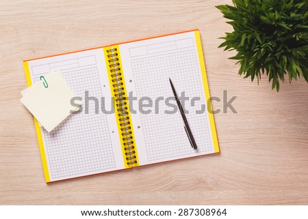 Opened notebook, pen, sticky notes and plant on bright wooden office desk.