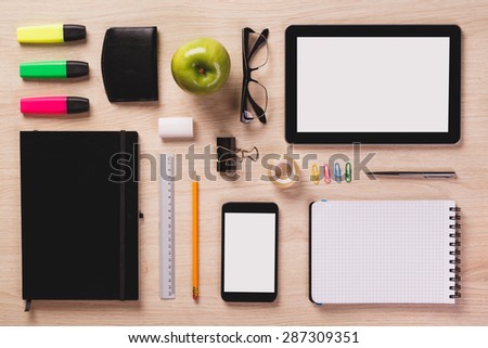 Digital tablet, smartphone, notepad, pen, pencil, rubber, green apple, calendar, highlighters and other office equipment arranged in perfect order on bright wooden office desk.