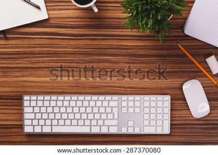 Blank space between modern white keyboard, mouse, pencil, mug of coffee, plant and other office equipment on dark wooden office desk.