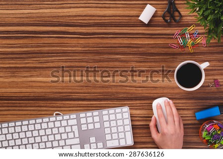 Someone is using computer keyboard and mouse next to modern black smartphone with empty screen, cup of black coffee and plant od dark wooden office desk.