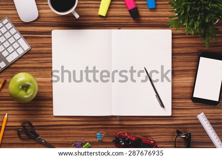Opened notebook in the middle of office equipment such as modern computer keyboard, mouse, apple, cup of coffee, highlighters, smartphone, plant, ruler and other on the dark wooden office desk.