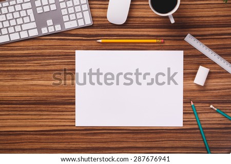 Blank sheet of paper next to pencil, eraser, ruler, modern computer keyboard, mouse and cup of coffee on dark wooden office desk.