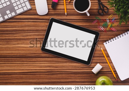 Digital modern black tablet with empty screen next to notepad, computer keyboard, mouse, pencil, cup of coffee, notepad, plant and other on dark wooden office table.