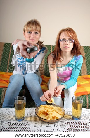 Two girls watching TV in living room, with two juice and some snack on table. Concept of entertainment, TV, friendship, relaxing