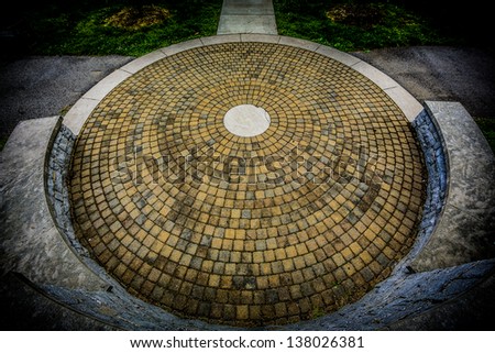 A circular stone floor with a round stone center piece surrounded by a raised stone wall and a concrete walking path