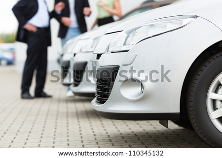 Two men man and one woman stand behind a row of silver-metallic cars on a parking place on yard of dealer