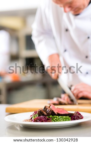 Chef in hotel or restaurant kitchen cooking, he is cutting meat or steak for a dish on plate