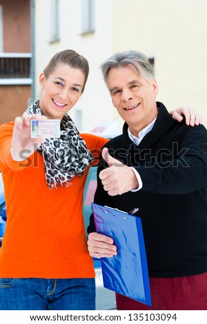 Driving school - Young woman has passed her driving test and proudly holding her driver license, the driving instructor is standing next to her