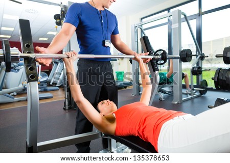 Woman with her personal fitness trainer in the gym exercising with dumbbells, she is using barbell on a weight bench