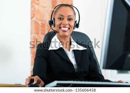 Young businesswoman or secretary working in her Office, she is sitting on the desk in front of the window and working on a computer with a headset, she has a customer pitch