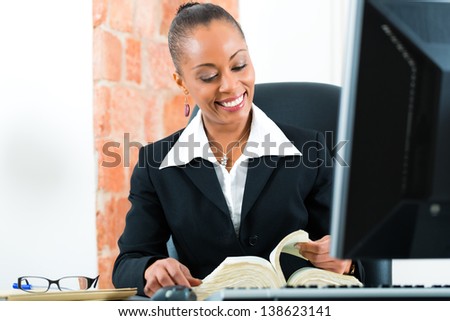 Young female black lawyer working in her office and reading a typical law book in front of a computer