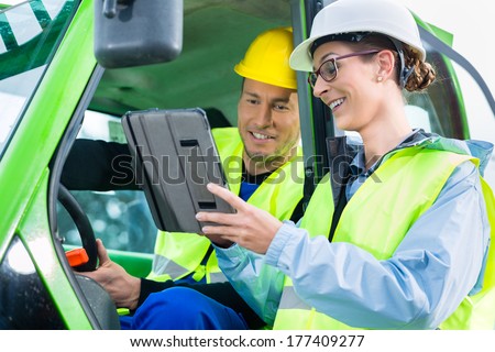 Construction worker in construction machinery discussing with engineer blueprints on pad or tablet computer on site