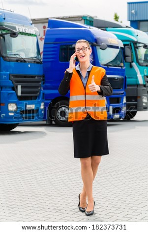 Logistics - female forwarder or supervisor with mobile phone, in front of trucks and trailers, on transshipment point
