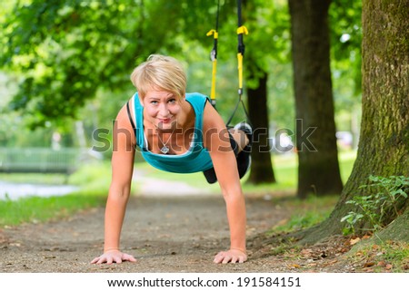 Young woman exercising with suspension trainer sling in City Park under summer trees for sport fitness