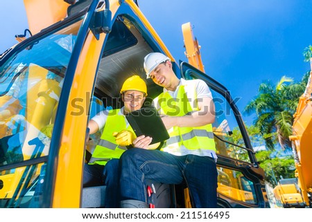Asian construction machinery driver discussing with foreman blueprints  or tablet computer of building site