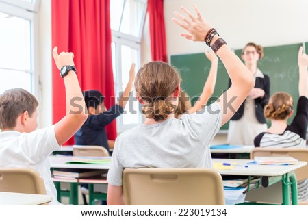 School class teacher giving lesson in front of a blackboard or board teaching students or pupils, they are raising their hands as they know all the answers