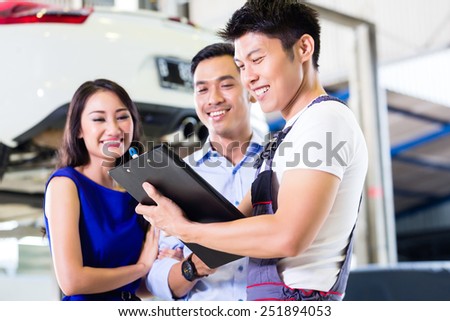 Car mechanic and Asian customer couple going through checklist with auto on hoist in the background of workshop